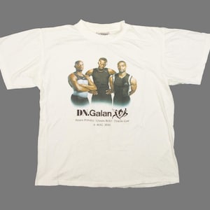 May include: White t-shirt featuring a graphic of three men in athletic attire. The text "DN.Galan" is printed below the image, along with names and the date "6 April 2008". The shirt is made of a soft, lightweight fabric.