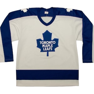 May include: A white hockey jersey with blue detailing and long sleeves. The jersey displays a blue maple leaf logo with the text "TORONTO MAPLE LEAFS". Blue stripes are on the sleeves and shoulders. The jersey is a classic design.