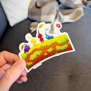 May include: A colorful sticker featuring cartoon characters with plant-like features. The sticker has a white border and a landscape design with green grass, a brown base, and various Pikmin characters in red, blue, yellow, purple, and white. The text "TSperring" is visible.