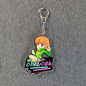 May include: A keyring with a cartoon character with orange hair and a green jacket. The character is sitting with their fingers in a peace sign. The keyring has a black base with the text "HP 192/084" in white, teal, and purple.
