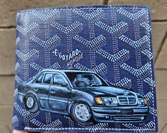 May include: A navy blue wallet featuring a repeating white pattern and a painted image of a black car. The wallet has the text "E. GOYARD HONORE" in white. The car has silver rims and a black body.