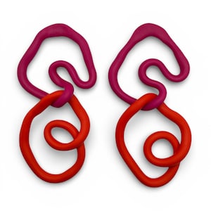 Könnte beinhalten: Ein Paar abstrakte Ohrringe in modernem Stil. Jeder Ohrring weist eine Kombination aus Formen und Farben auf. Der obere Teil ist ein tiefes Magenta, verbunden mit einer leuchtend roten Schlaufe mit Spiraldesign. Die Ohrringe sind vor weißem Hintergrund.