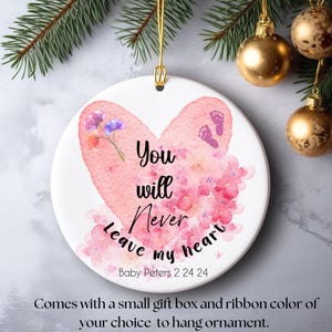 Puede incluir: Un adorno de cerámica rosa y blanco en forma de corazón con el texto "You will never leave my heart" y el nombre "Baby Peters 2 24 24" debajo. El adorno está decorado con flores rosas y un par de huellas de pies de bebé.