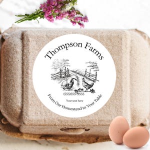 May include: A carton of eggs with a round label that reads "Thompson Farms." The label features a black and white illustration of chickens and a road. The text "From Our Homestead to Your Table" is also on the label. Two brown eggs are in the foreground.