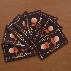 May include: A collection of playing cards fanned out on a wooden surface. Each card features a dark brown background with an acorn and leaf design in shades of orange, yellow, and white, framed by a decorative border.