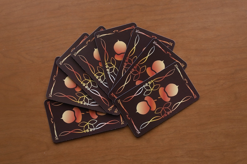 May include: A collection of playing cards fanned out on a wooden surface. Each card features a dark brown background with an acorn and leaf design in shades of orange, yellow, and white, framed by a decorative border.
