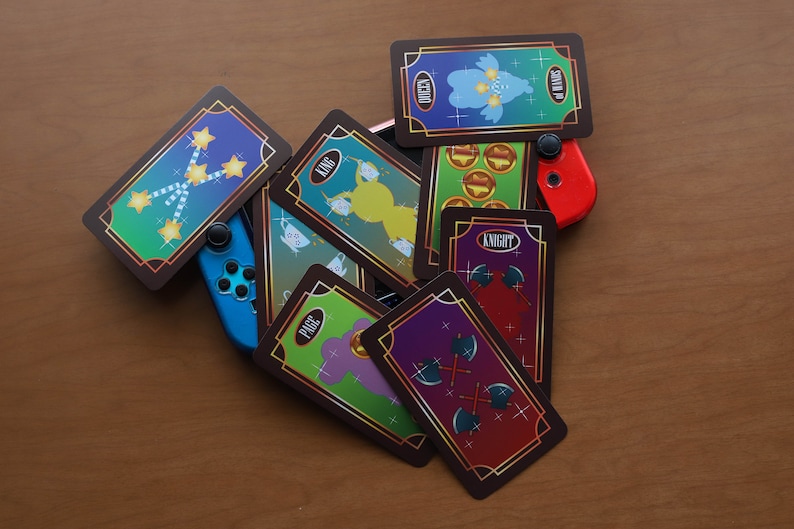 May include: A collection of illustrated playing cards with gold trim, featuring various characters and symbols, are arranged with blue and red game controllers on a wooden surface. The cards display images of stars, coins, and axes.