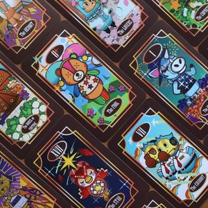 May include: A collection of tarot cards with brown borders and gold accents. Each card features colorful illustrations of cartoon characters and various symbols. The cards are arranged at different angles, showcasing the detailed artwork and text labels.