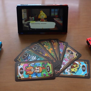 May include: A Nintendo Switch console with a screen displaying a video game scene. Two Joy-Con controllers, one blue and one red, flank a deck of illustrated cards. The game screen shows the text "Good morning, everyone!"