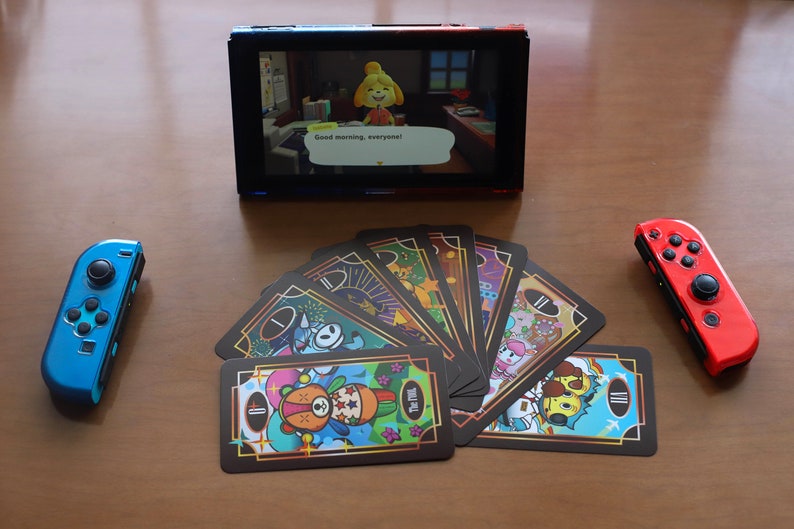 May include: A Nintendo Switch console with a screen displaying a video game scene. Two Joy-Con controllers, one blue and one red, flank a deck of illustrated cards. The game screen shows the text "Good morning, everyone!"