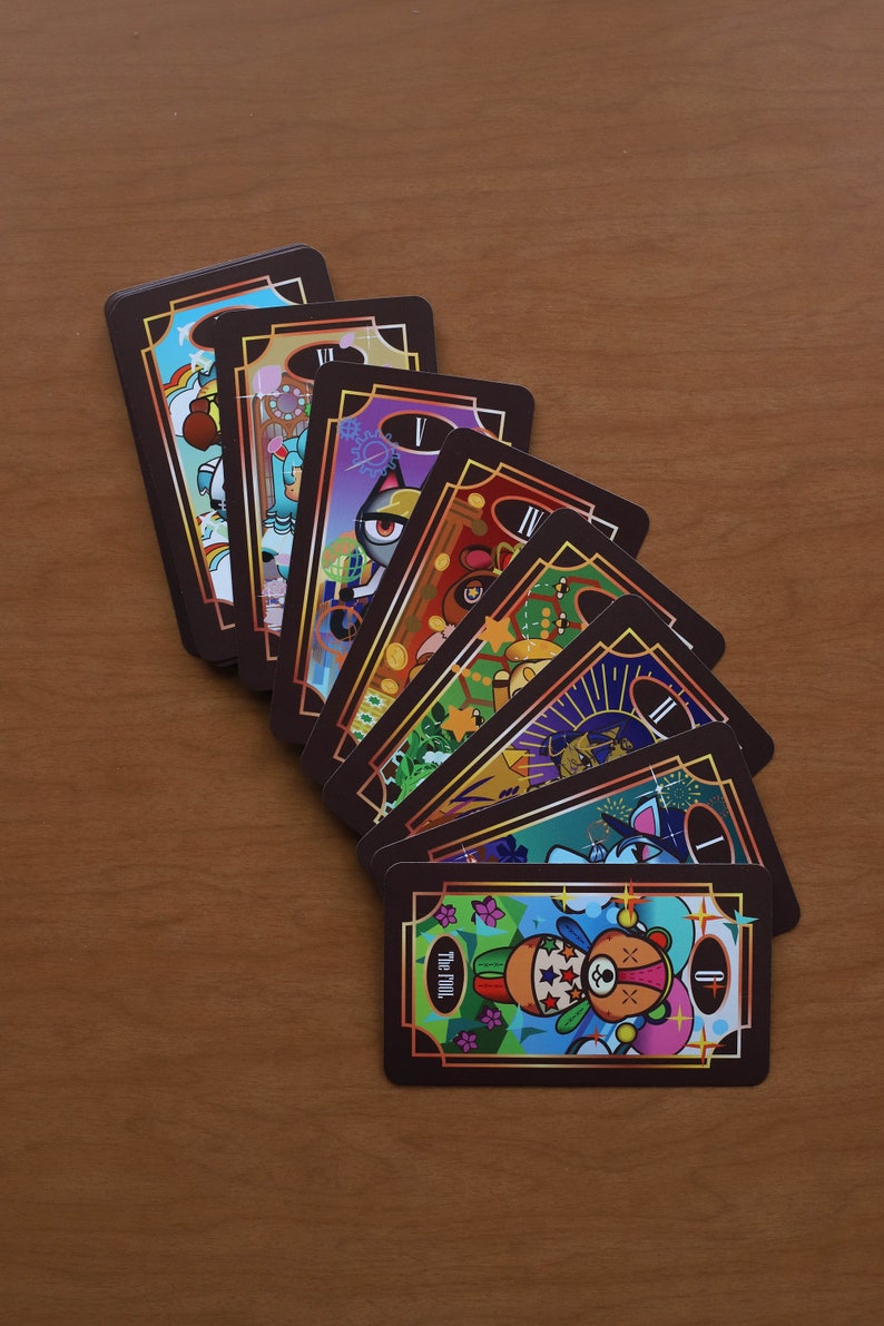 May include: A fan of tarot cards with colorful illustrations and gold accents. The cards feature various whimsical designs, including a cat with a gear eye, a bear, and other cartoon characters. The cards are arranged on a wooden surface.