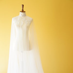 Könnte beinhalten: Eine weiße Tüll-Hochzeitscape mit hohem Kragen und zarten Stickereien. Die Cape ist über eine Kleiderpuppe drapiert und hat einen langen Schleier.