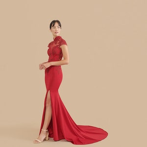 Pode incluir: Uma mulher vestindo um vestido vermelho longo at&eacute; o ch&atilde;o, com gola alta e uma fenda lateral. O vestido tem detalhes delicados de renda no corpete e uma cauda.