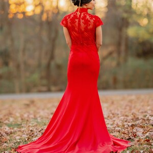 Pode incluir: Uma mulher vestindo um vestido vermelho de renda, longo at&eacute; o ch&atilde;o e com uma cauda longa. Ela est&aacute; em p&eacute; em um campo de folhas ca&iacute;das.