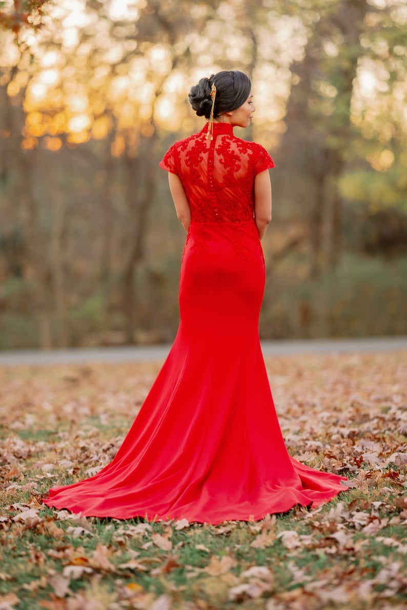 Pode incluir: Uma mulher vestindo um vestido vermelho de renda, longo at&eacute; o ch&atilde;o e com uma cauda longa. Ela est&aacute; em p&eacute; em um campo de folhas ca&iacute;das.