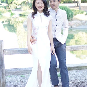 May include: A couple poses for a photo in front of a wooden fence. The woman is wearing a white dress with a lace overlay and a high slit. The man is wearing a light gray button-down shirt with a beige vest and dark gray pants.