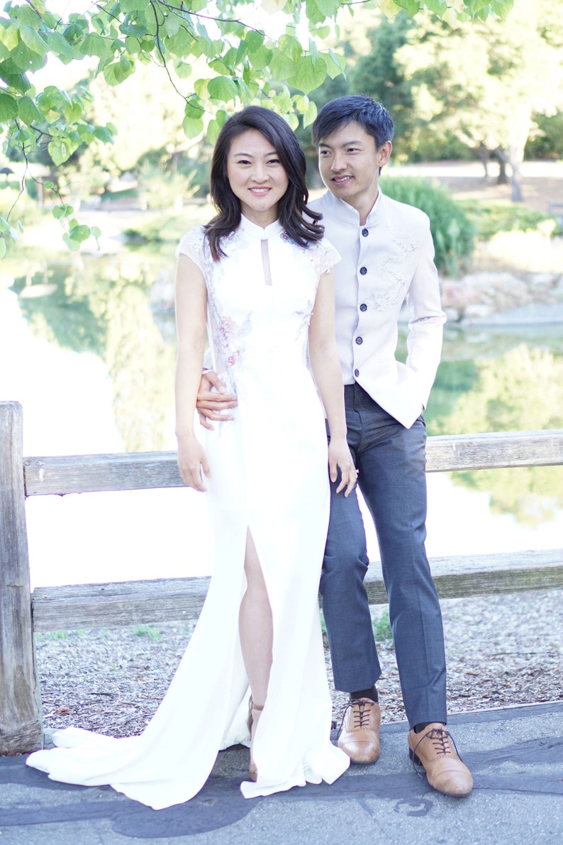 May include: A couple poses for a photo in front of a wooden fence. The woman is wearing a white dress with a lace overlay and a high slit. The man is wearing a light gray button-down shirt with a beige vest and dark gray pants.