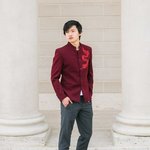 May include: A man wearing a burgundy blazer with a red dragon embroidered on the front, gray pants, and brown leather shoes. He is standing in front of a white wall with pillars.