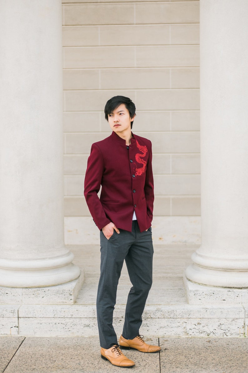 May include: A man wearing a burgundy blazer with a red dragon embroidered on the front, gray pants, and brown leather shoes. He is standing in front of a white wall with pillars.