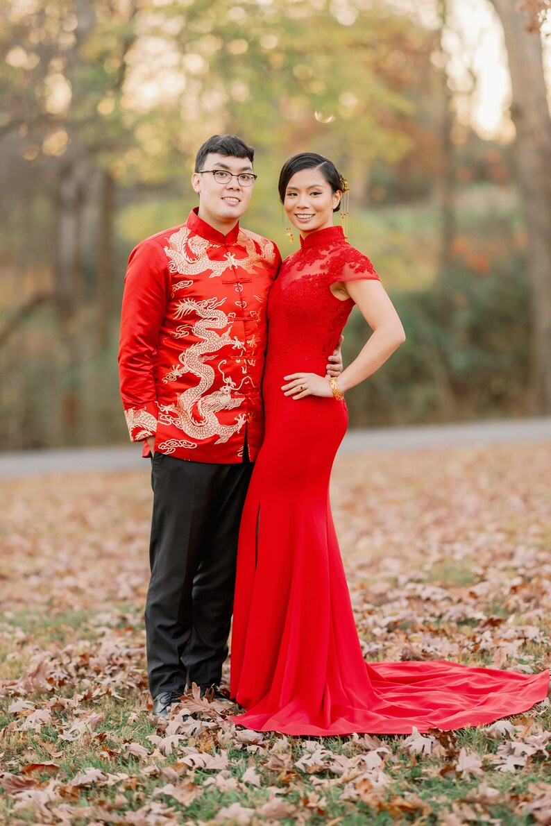 Pode incluir: Um casal com trajes tradicionais de casamento chin&ecirc;s. O homem est&aacute; vestindo uma jaqueta vermelha com bordados de drag&otilde;es dourados e cal&ccedil;as pretas. A mulher est&aacute; vestindo um vestido vermelho com gola alta e uma cauda longa. Eles est&atilde;o em p&eacute; em um campo de folhas ca&iacute;das.