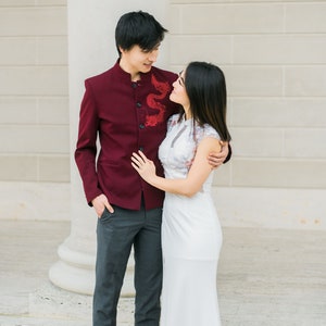 May include: A couple in traditional Chinese wedding attire. The man is wearing a burgundy jacket with a dragon embroidered on the front and gray pants. The woman is wearing a white dress with lace detailing. They are standing in front of a white pillar and looking at each other.