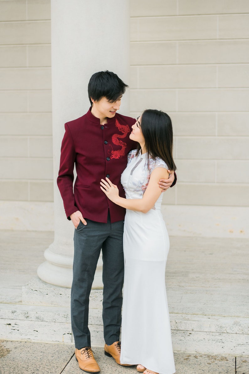 May include: A couple in traditional Chinese wedding attire. The man is wearing a burgundy jacket with a dragon embroidered on the front and gray pants. The woman is wearing a white dress with lace detailing. They are standing in front of a white pillar and looking at each other.