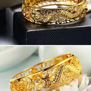 May include: A gold-colored bracelet with a dragon design. The bracelet is made of a shiny, metallic material and features intricate cut-out details. The dragon is a prominent feature, with detailed scales and a flowing design. The bracelet is a cuff style.