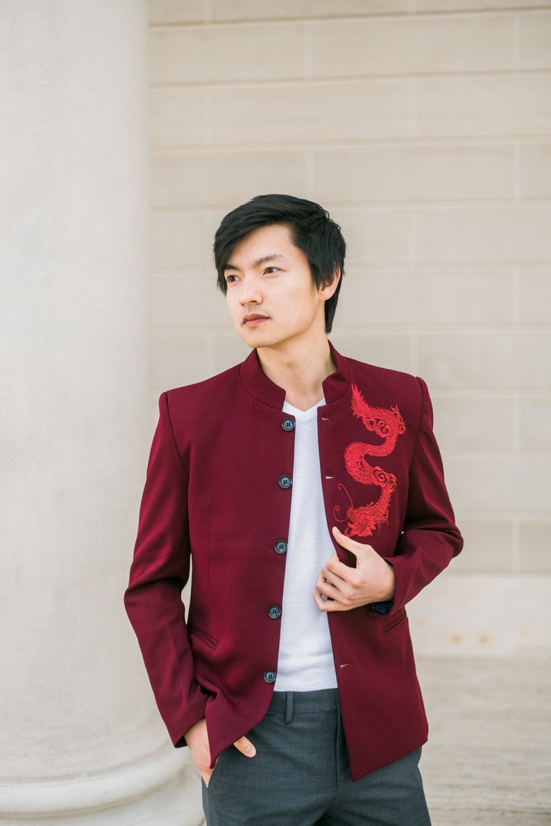May include: A man wearing a burgundy blazer with a red embroidered dragon design. The blazer has a mandarin collar and five black buttons. He is also wearing a white t-shirt and gray pants.