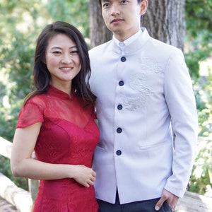 May include: A couple in traditional Chinese wedding attire. The woman is wearing a red dress with lace detailing and the man is wearing a white jacket with embroidered dragons and dark pants.