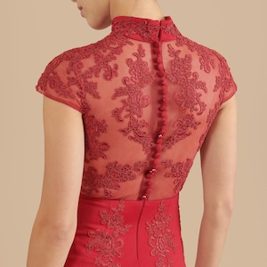 Pode incluir: Um vestido vermelho com uma sobreposi&ccedil;&atilde;o de renda transparente e uma gola alta. O vestido tem uma silhueta ajustada e apresenta uma fila de bot&otilde;es nas costas.
