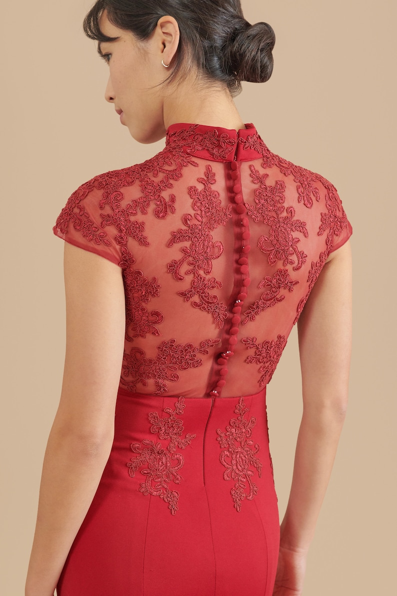 Pode incluir: Um vestido vermelho com uma sobreposi&ccedil;&atilde;o de renda transparente e uma gola alta. O vestido tem uma silhueta ajustada e apresenta uma fila de bot&otilde;es nas costas.