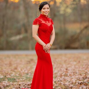 Pode incluir: Uma mulher com um vestido vermelho longo de renda, de manga comprida e gola alta. Ela est&aacute; em p&eacute; em um campo de folhas ca&iacute;das.