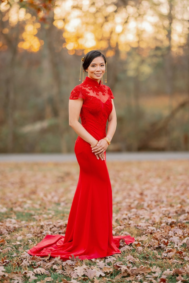Pode incluir: Uma mulher com um vestido vermelho longo de renda, de manga comprida e gola alta. Ela est&aacute; em p&eacute; em um campo de folhas ca&iacute;das.
