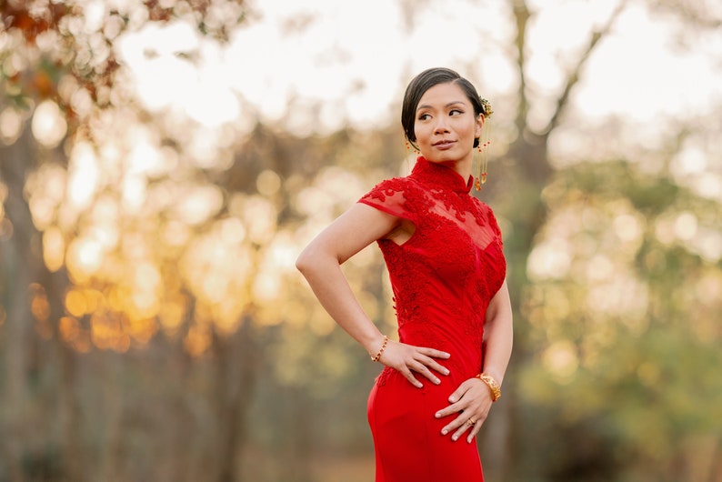 Pode incluir: Uma mulher com um vestido vermelho com bordados intrincados est&aacute; em um ambiente de floresta. O vestido tem um colar alto e mangas curtas. A mulher est&aacute; olhando para o lado, com o cabelo puxado para tr&aacute;s.