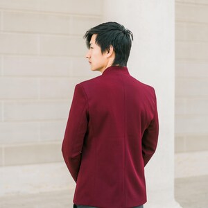 May include: A man wearing a burgundy blazer with a mandarin collar and gray pants, standing in front of a white wall.