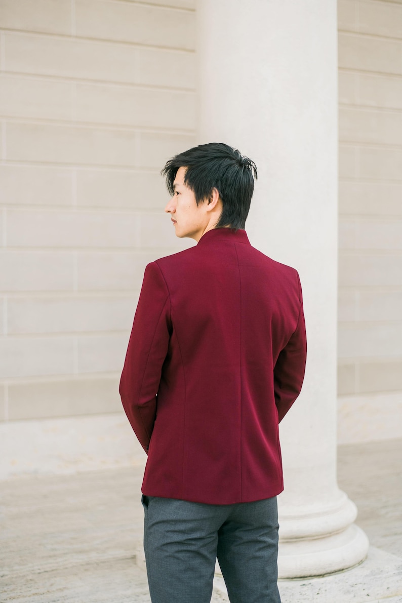 May include: A man wearing a burgundy blazer with a mandarin collar and gray pants, standing in front of a white wall.