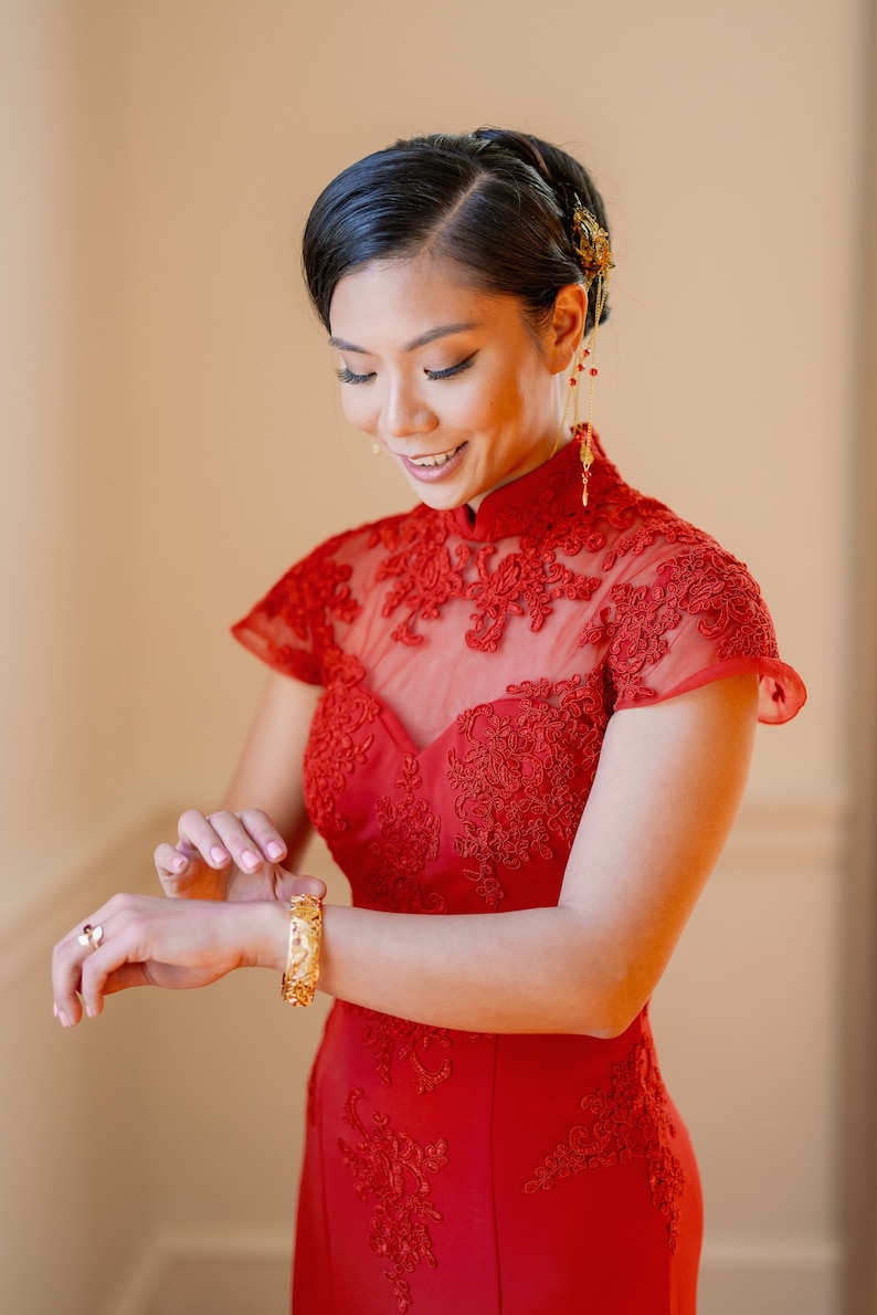Pode incluir: Uma mulher com um vestido vermelho com gola alta e mangas curtas, adornado com renda. Ela est&aacute; usando joias de ouro e tem o cabelo puxado para tr&aacute;s.