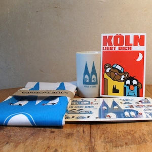 Puede incluir: Un conjunto de regalos con temática de Colonia que incluye un paño de cocina azul y blanco con el texto "Vorsicht Koln", una taza blanca con el texto "Kölle is cool" y una ilustración azul y blanca de una iglesia, una tarjeta roja con el texto "Köln liebt dich" y una ilustración azul y blanca de un oso y un hombre, y un posavasos azul y blanco con el texto "Kölle ist cool" y una ilustración azul y blanca de una iglesia.