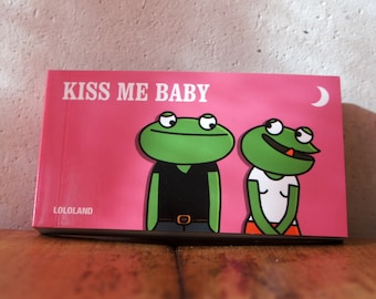 May include: A pink book cover with the text "KISS ME BABY" and two cartoon frogs. The frogs are green with black outlines, one wearing a black shirt and the other a white shirt. A crescent moon is in the upper right corner.