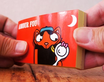 May include: A red and yellow notepad with a cartoon cat wearing a blue hat and holding a white object. The notepad has the text "AMOUR FOU" on the front.