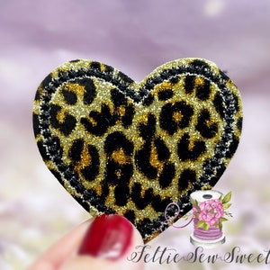 May include: A heart-shaped patch with a leopard print design. The patch features a black border with gold glitter accents. The leopard print is a mix of black and gold, creating a sparkling effect. The background is a soft purple.