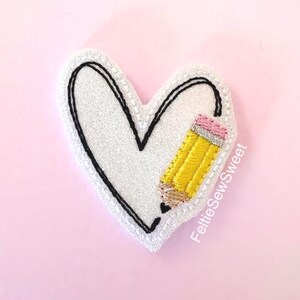 May include: A white heart-shaped patch with a glittery texture, featuring a black embroidered outline and a yellow pencil with a pink eraser and silver ferrule. The text FeltieSewSweet is embroidered in white along the right side of the heart.