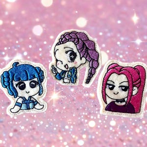 May include: Three embroidered patches featuring anime-style characters with large eyes and detailed hair. One has blue hair, another purple, and the third has pink hair. Each patch is outlined in white and set against a pink glitter background.