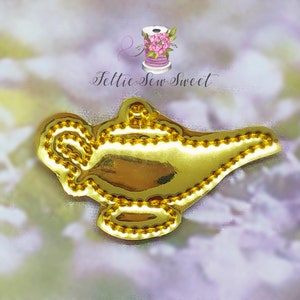 May include: A gold-colored genie lamp-shaped applique with a beaded border. The applique is shiny and reflective, with a detailed design. The background is a soft, blurred mix of purple, pink, and green hues. The text "Feltie Sew Sweet" is at the top.