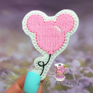 May include: Embroidered patch in the shape of a pink Mickey Mouse balloon. The balloon has a white outline and a black string. The patch is on a blurred background. The text "Feltie Sew Sweet" is visible.
