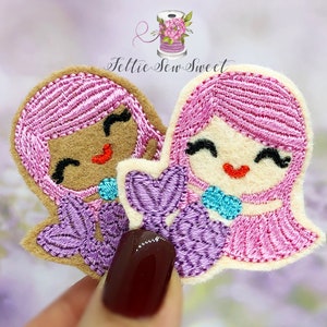 May include: Two felt mermaid appliques with pink hair, purple tails, and smiling faces. One mermaid has brown skin and the other has light skin.