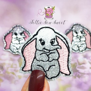 May include: Three silver glitter embroidered iron-on patches of Dumbo the elephant with pink ears.