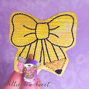 May include: A yellow bow with black outlines and a pencil design. The bow is made of a sparkly fabric.