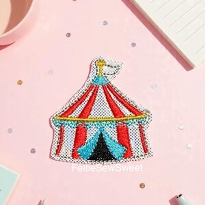 May include: Embroidered patch of a circus tent with red and white striped roof, blue and black entrance, and a gold flag. The patch has a white mesh background and is on a pink surface. The text "FeltieSewSweet" is at the bottom.