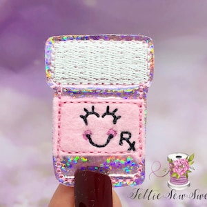 May include: Pink felt pill bottle with a smiling face and the letter "R" embroidered on it. The bottle is decorated with iridescent glitter.
